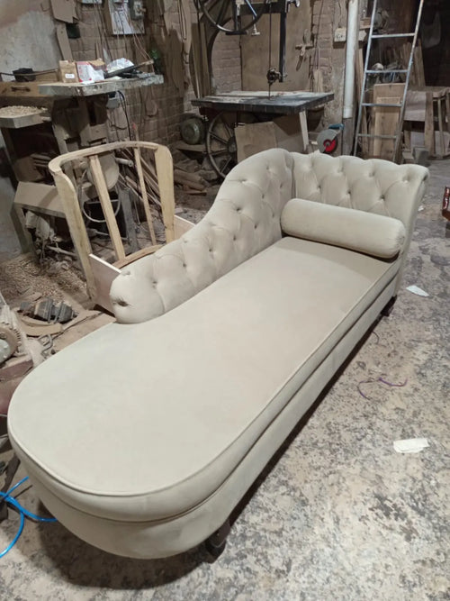 A tufted chaise lounge daybed in a light beige fabric, photographed in a cluttered furniture workshop.