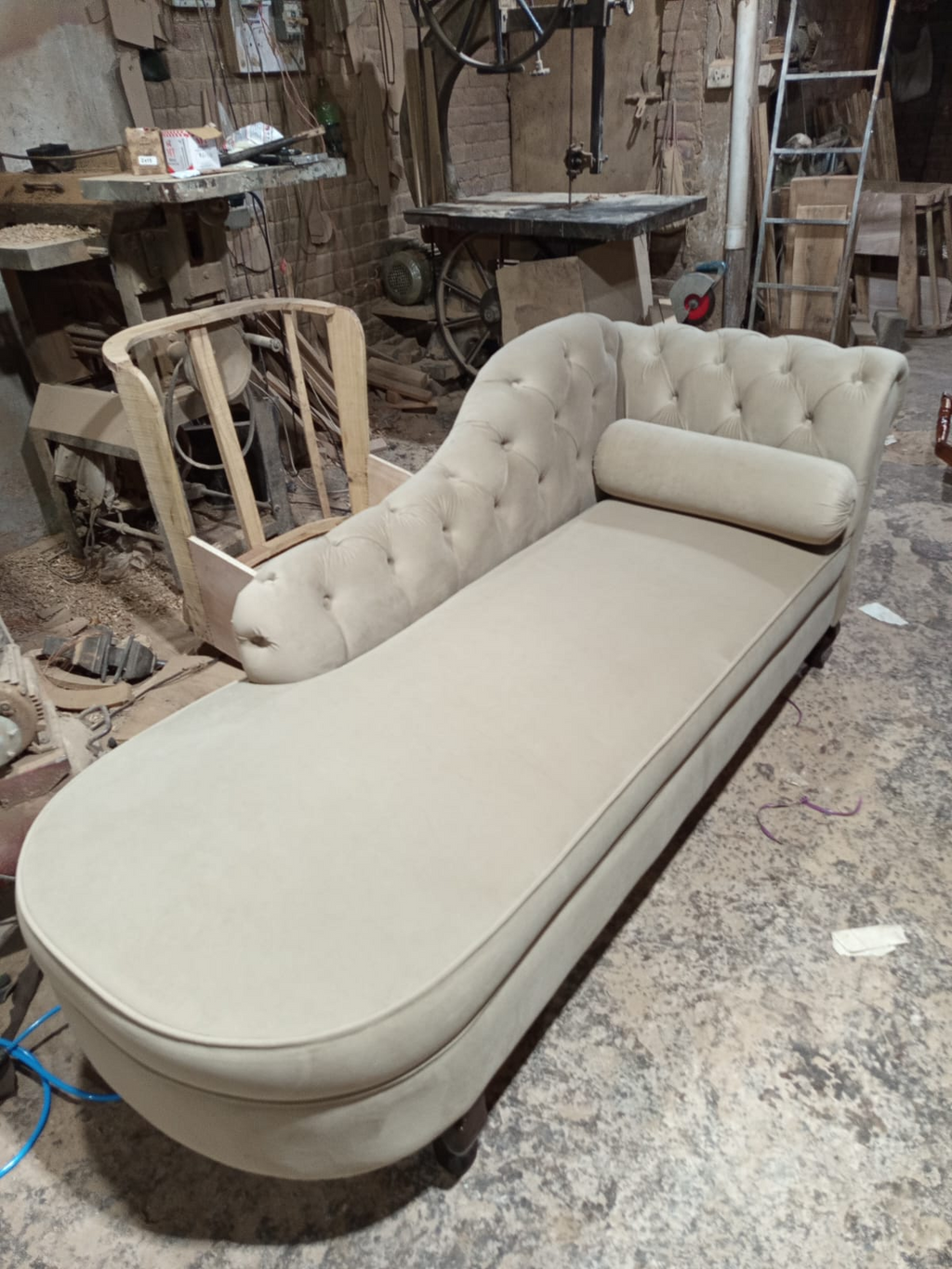 A tufted chaise lounge daybed in a light beige fabric, photographed in a cluttered furniture workshop.