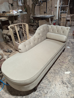 A tufted chaise lounge daybed in a light beige fabric, photographed in a cluttered furniture workshop.