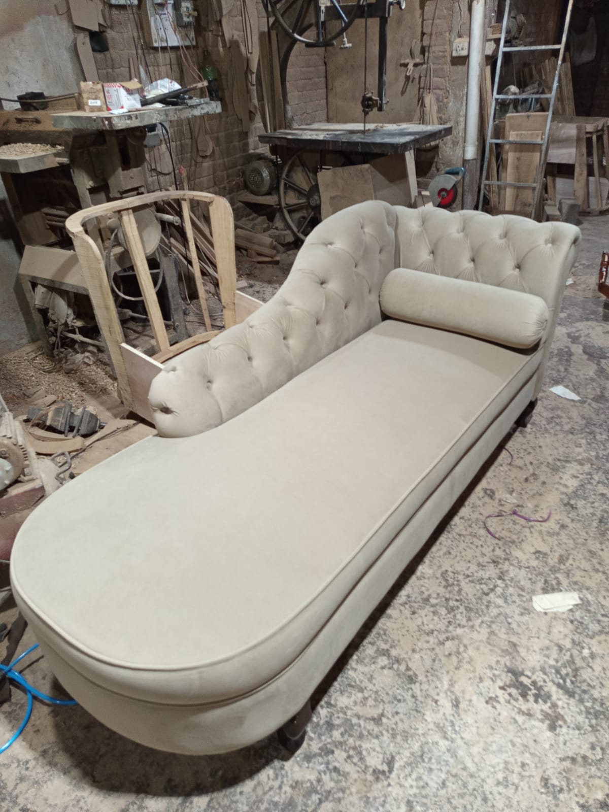 A tufted chaise lounge daybed in a light beige fabric, photographed in a cluttered furniture workshop.
