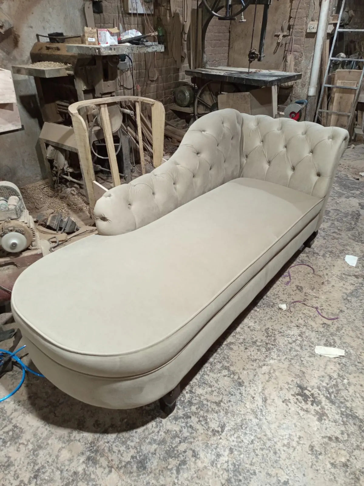 A tufted chaise lounge chaise lounge daybed upholstered in a light beige fabric, photographed in a cluttered furniture workshop.