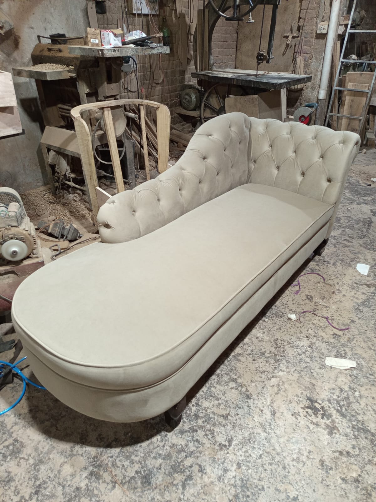 A tufted chaise lounge chaise lounge daybed upholstered in a light beige fabric, photographed in a cluttered furniture workshop.