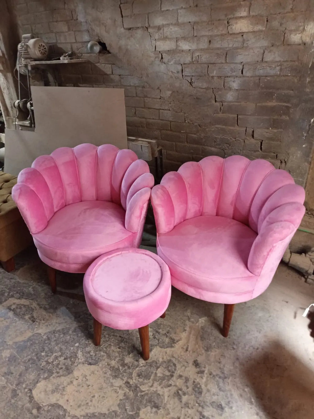 A pair of pink velvet shell-back armchairs with a matching round ottoman, set against a rustic brick and concrete wall background.