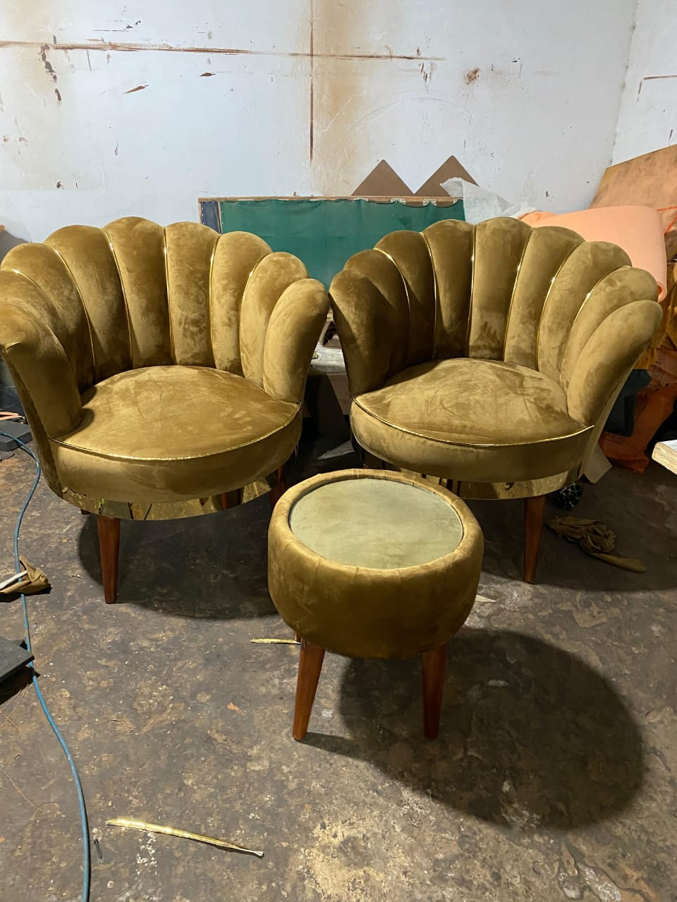 A pair of olive green velvet armchairs with a scalloped shell back design and a matching round ottoman, set in a workshop environment.