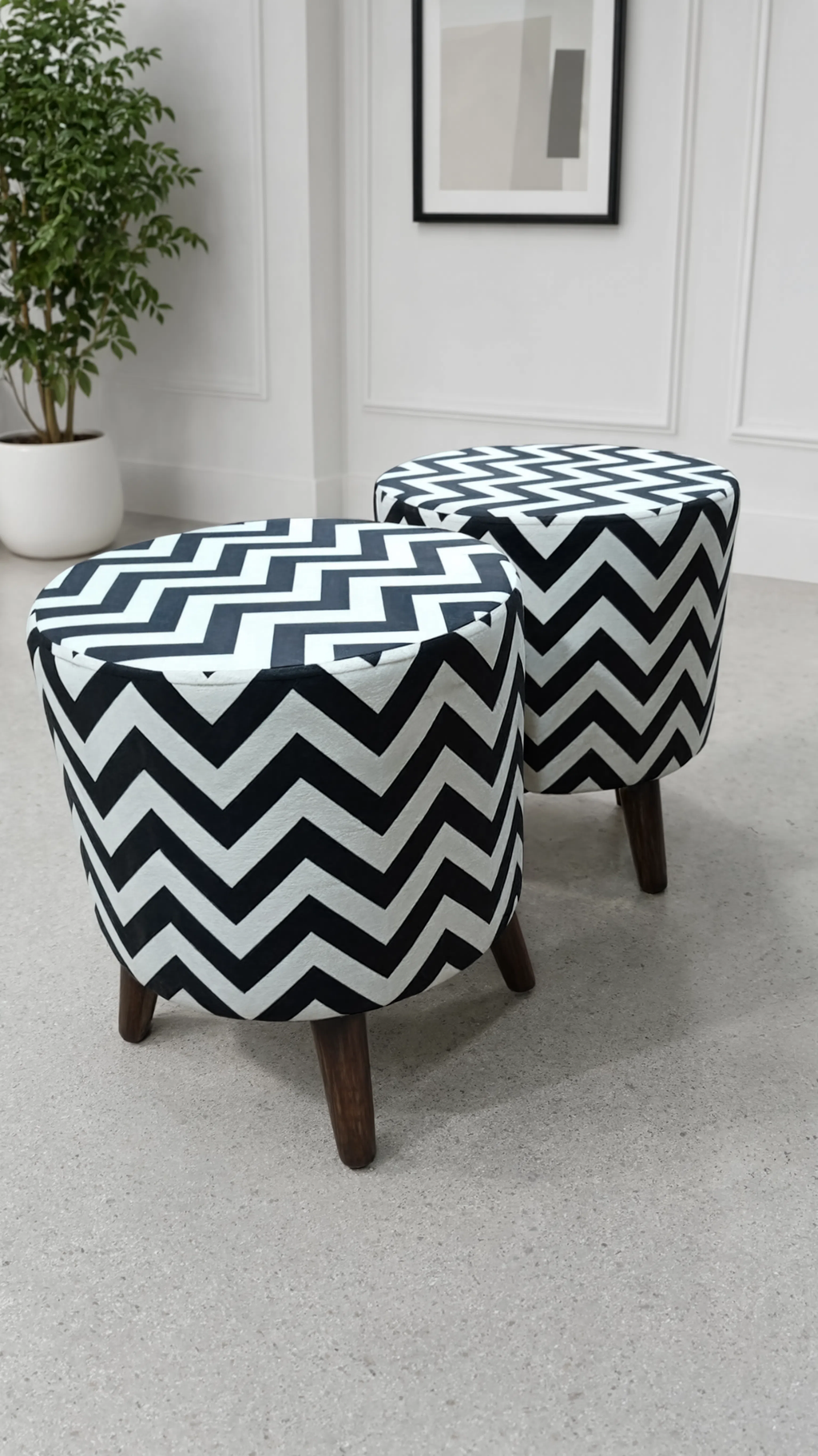 Two round upholstered ottomans with a bold black and white chevron pattern and dark wooden legs, shown in a bright room with a potted plant and framed wall art.