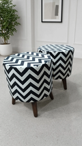 Two round upholstered ottomans with a bold black and white chevron pattern and dark wooden legs, shown in a bright room with a potted plant and framed wall art.