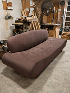 A curved, tufted daybed in a brown bouclé fabric, photographed in a cluttered workshop environment with wood panels, tools, and in a workshop.