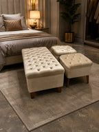 A tufted cream velvet storage bench and two matching ottomans styled in a modern, warm-toned bedroom setting on a gray rug.