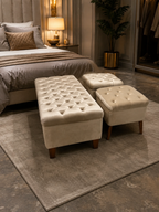 A tufted cream velvet storage bench and two matching ottomans styled in a modern, warm-toned bedroom setting on a gray rug.