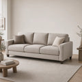 A light beige upholstered three-seat sofa with two matching throw pillows, shown in a minimalist living room setting with a wooden coffee table, a large neutral rug, and soft natural light.
