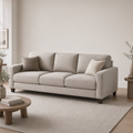 A light beige upholstered three-seat sofa with two matching throw pillows, shown in a minimalist living room setting with a wooden coffee table, a large neutral rug, and soft natural light.