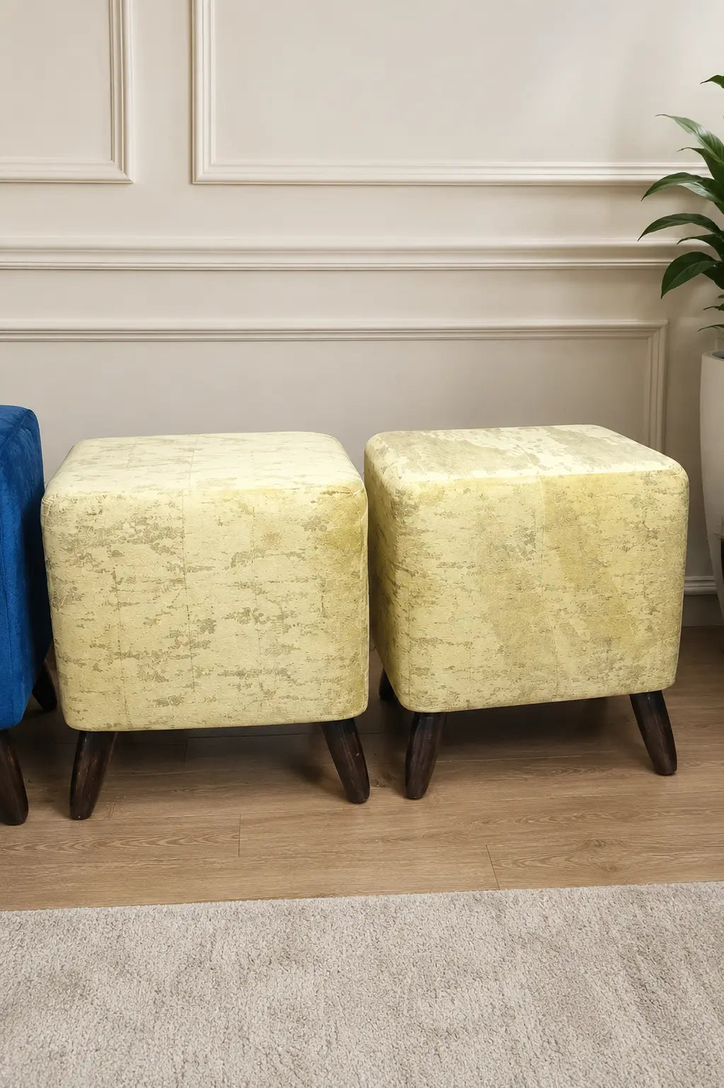 Two square ottomans with textured pale yellow upholstery and dark wooden legs, shown in an indoor room setting with a paneled wall, wood floor, and a plant.