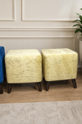 Two square ottomans with textured pale yellow upholstery and dark wooden legs, shown in an indoor room setting with a paneled wall, wood floor, and a plant.