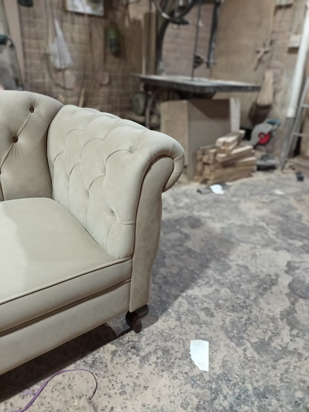 A close-up of a beige tufted armchair in a workshop or furniture shop setting with a concrete floor and workbench in the background.