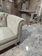 A close-up of a beige tufted armchair in a workshop or furniture shop setting with a concrete floor and workbench in the background.