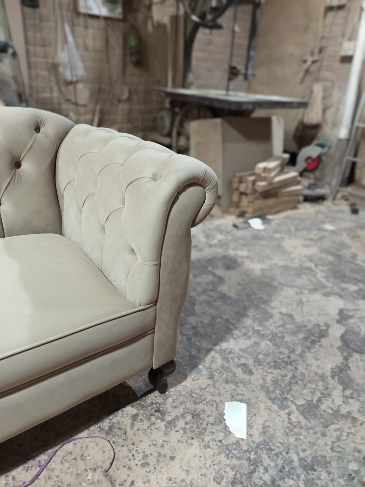 A close-up of a beige tufted armchair in a workshop or furniture shop setting with a concrete floor and workbench in the background.