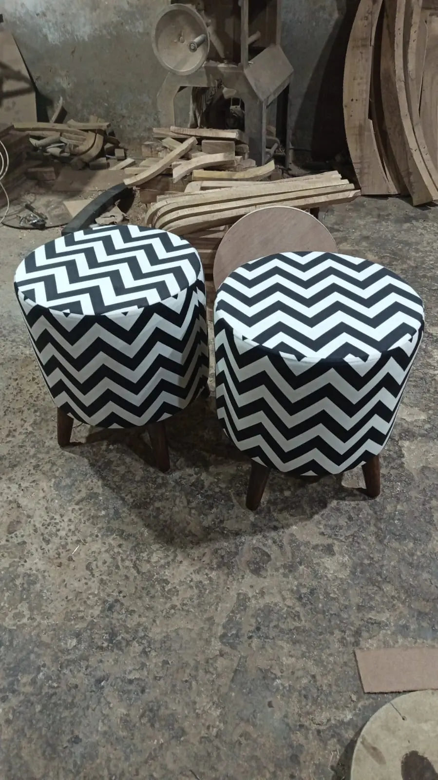 Two round upholstered stools with black and white chevron-patterned fabric and dark wooden legs, photographed in a workshop or furniture shop with wood scraps and machinery in the background.