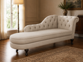 An upholstered chaise lounge in a warm off-white fabric, featuring tufted button detailing on the backrest and seat, supported by dark turned wooden legs, set in a classic room with hardwood floors and a patterned rug.