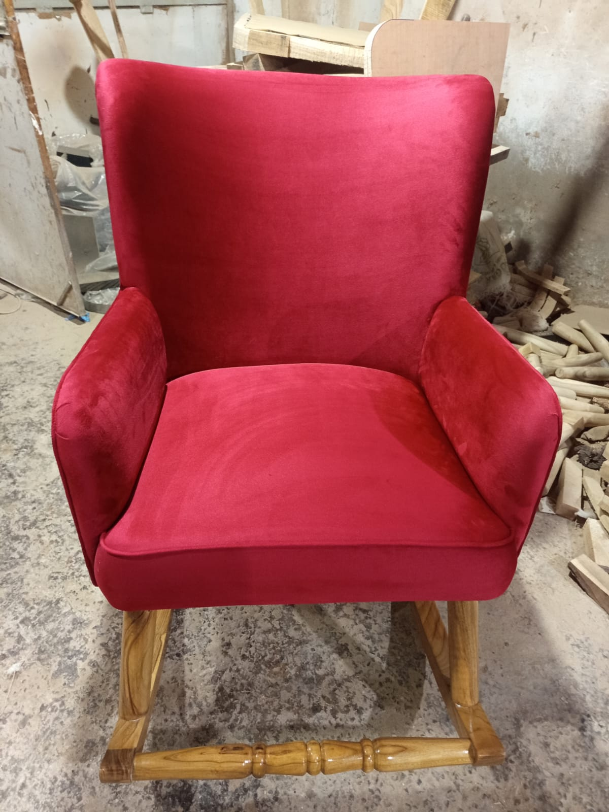 A red velvet upholstered rocking chair with a natural wood frame, photographed in a workshop or furniture shop setting.