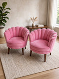 Two pink shell-back velvet armchairs with dark wooden legs, placed on a woven beige rug in a bright, modern room with a plant and a small wooden side table in the background.