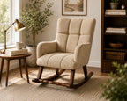 A beige upholstered rocking chair with a high back and tufted seat, shown in a styled living room setting with a side table, lamp, plant, rug, and bookshelf.