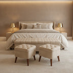 A staged bedroom scene with a bed dressed in cream bedding, two matching upholstered ottomans at the foot of the bed, side tables with lit lamps, and a warm beige wall and carpet.