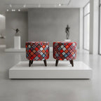 Two small upholstered stools with dark wooden legs sit side by side on a white display platform in a modern gallery space. The stools feature a dense geometric patchwork pattern in red, blue, orange, yellow, and white.