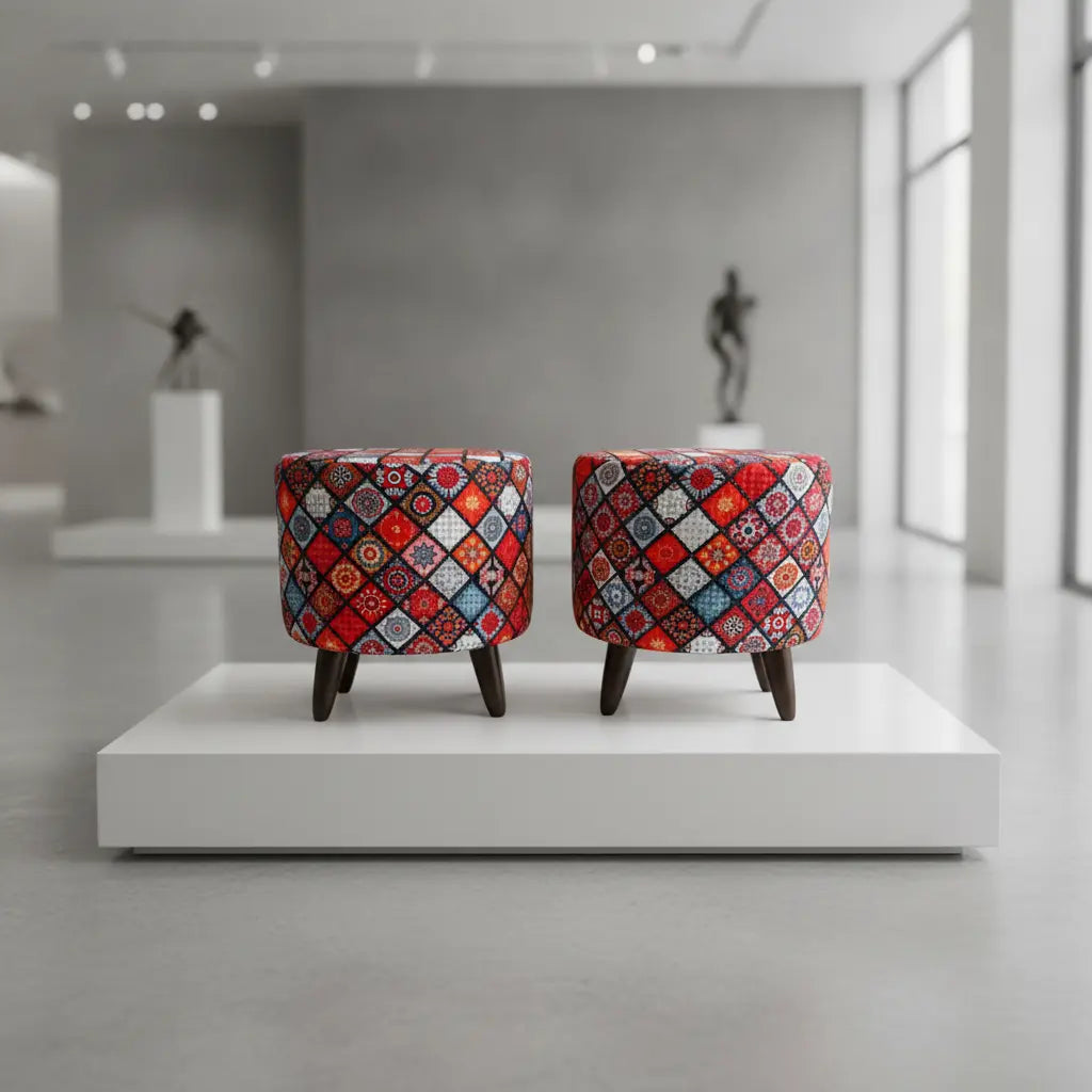 Two small upholstered stools with dark wooden legs sit side by side on a white display platform in a modern gallery space. The stools feature a dense geometric patchwork pattern in red, blue, orange, yellow, and white.