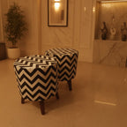 Two round upholstered ottomans with a bold black and cream chevron pattern and dark wooden legs, placed in a warm-toned indoor room with marble walls and a potted plant.