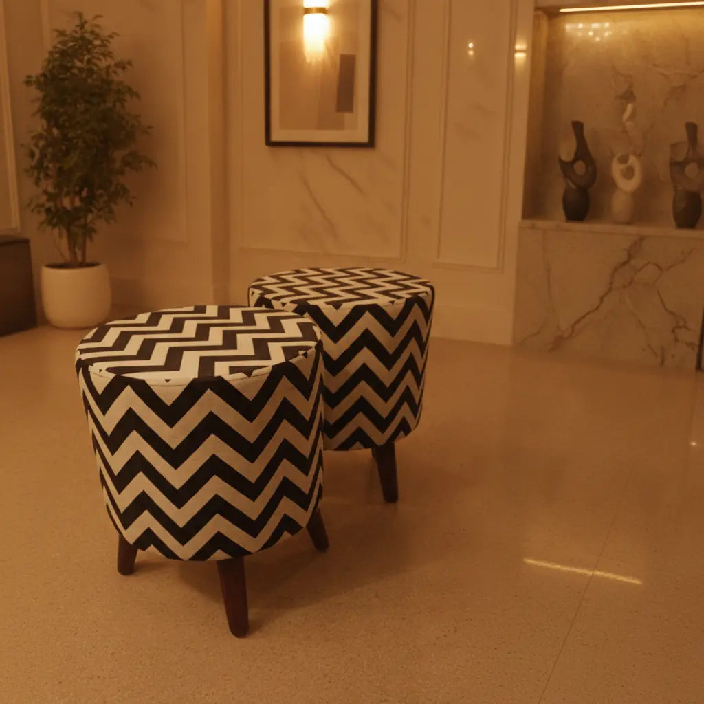 Two round upholstered ottomans with a bold black and cream chevron pattern and dark wooden legs, placed in a warm-toned indoor room with marble walls and a potted plant.