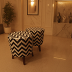 Two round upholstered ottomans with a bold black and cream chevron pattern and dark wooden legs, placed in a warm-toned indoor room with marble walls and a potted plant.