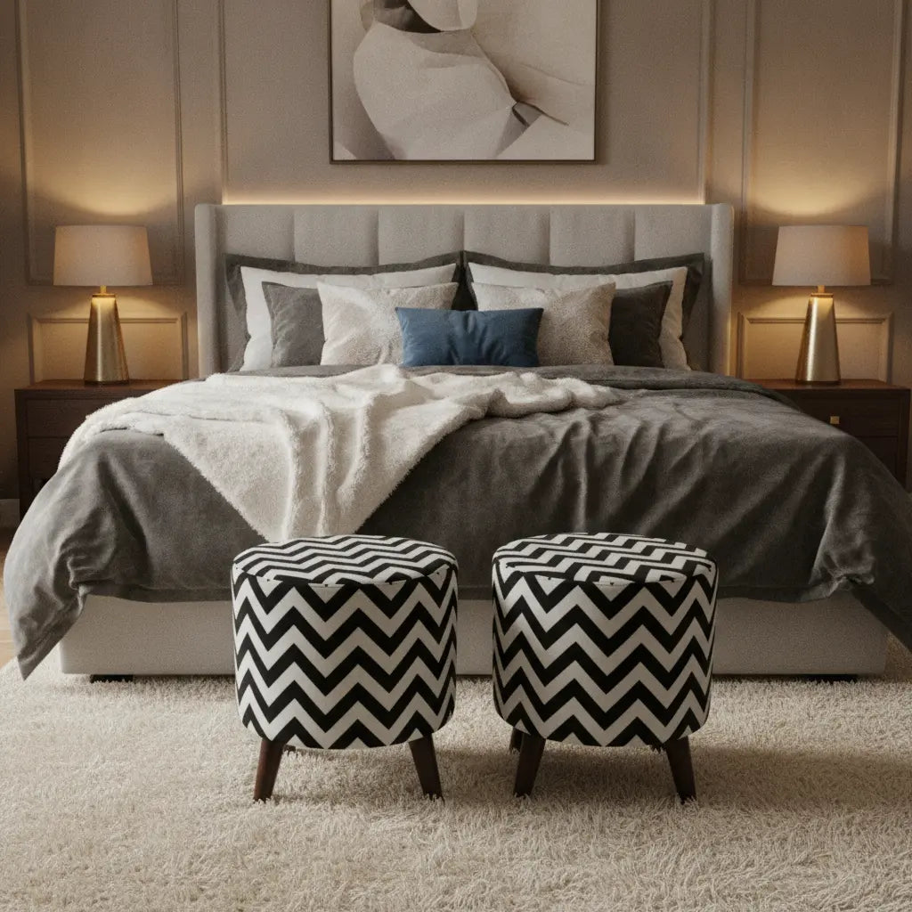 A styled bedroom scene with a bed dressed in gray and white bedding, two matching black-and-white chevron pouf ottomans at the foot of the bed, bedside tables with lit lamps, and a neutral wall with framed wall art.