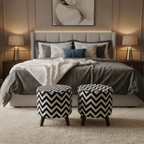 A styled bedroom scene with a bed dressed in gray and white bedding, two matching black-and-white chevron pouf ottomans at the foot of the bed, bedside tables with lit lamps, and a neutral wall with framed wall art.